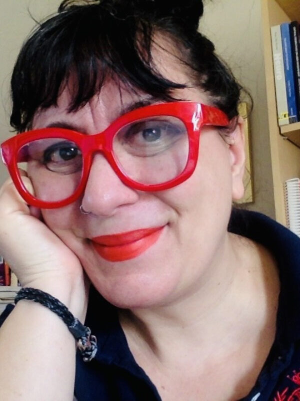 A woman with black hair and bangs, wearing bright red thick-rimmed glasses and bright red lipstick, smiling and leaning her head on the heel of her right hand.