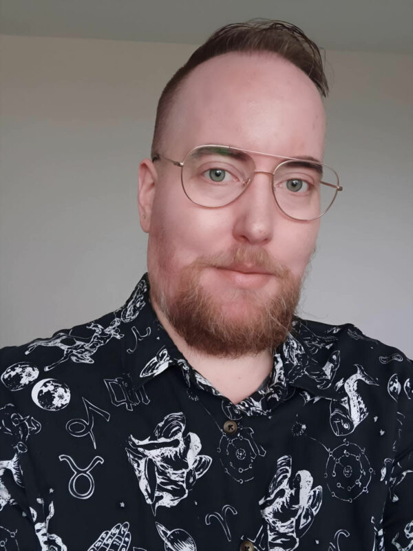 A person with short red hair and a trimmed red beard, wearing glasses and thinly smiling. He is wearing a black shirt with white abstract motifs on it.